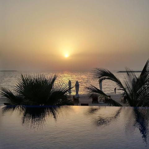 A photo of an infinity pool next to a beach and the sea at sunset. The sun is setting over the sea, casting a warm glow on the water. Palm trees and beach umbrellas line the shore.