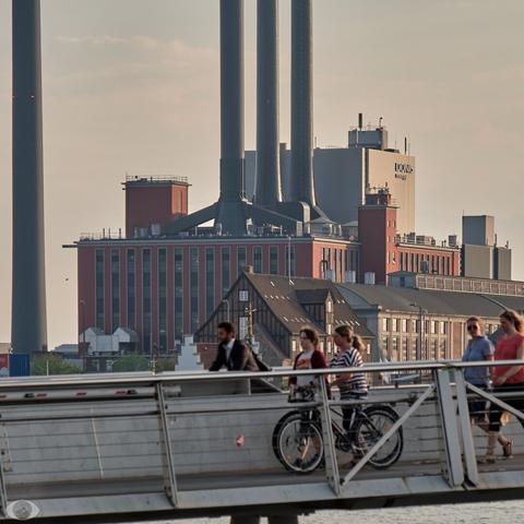 Rectangular brick building with large windows and 3 tall chimneys next to each other. On the left is an even taller and isolated chimney. In the foreground (blurred) is a cycle bridge with people cycling and walking.