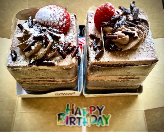 Two slices of Black Forest Cake with chocolate shavings coated with powdery frosting and strawberry on top sitting in little paper boxes.