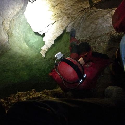 View down between the muddy trouser-legs and boots of a person. Another person in caving suit, helmet and hedalamp looing at a small clear pool between very rugged limestone rocks.