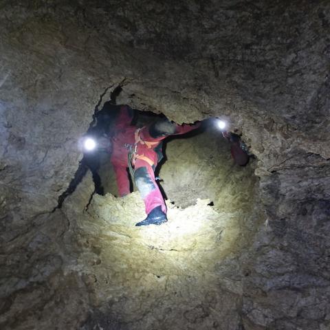 View from below through a hole in rugged limestone rocks, a person in caving gear traversing over the rock looking down at the camera.