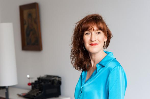 Das Bild zeigt eine Frau mit rötlich-braunem, lockigem Haar, die ein leuchtend blaues, seidiges Hemd trägt. Sie hat rote Lippen und trägt dezente Ohrringe. Ihr Blick ist freundlich und direkt in die Kamera gerichtet. Im Hintergrund, etwas unscharf, sind ein Gemälde mit einem goldenen Rahmen und eine alte Schreibmaschine zu erkennen. Das Licht ist weich und gleichmäßig, was eine angenehme Atmosphäre erzeugt. Der Hintergrund ist hellgrau, was die Farben der Frau und ihrer Kleidung hervorhebt. Insgesamt wirkt das Bild warm und einladend, mit einem Hauch von Vintage durch die Schreibmaschine im Hintergrund.