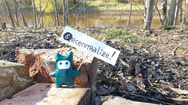 A photograph of a small blue figure with a mask, holding a little "decentralized" handwritten banner with "d" being the delta logo symbol.  Background of the photo is a forest and a lake in early spring.