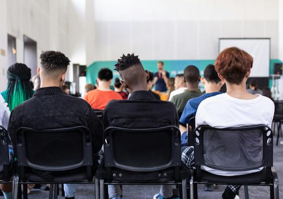 Ein Schulungsraum, in dem sitzende Personen in Stuhlreihen zu sehen sind. Sie sind mit den Rücken zugewandt.