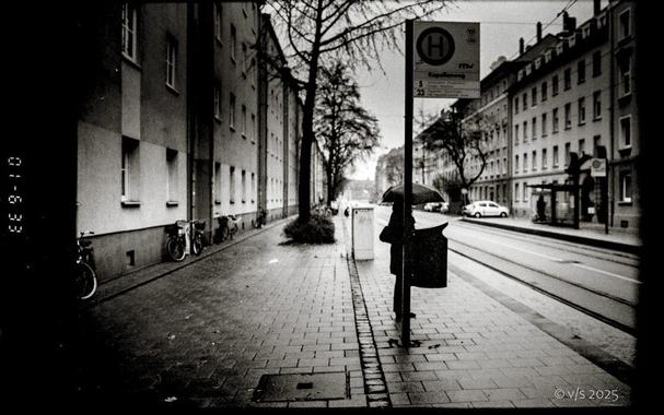 Eine Person mit Regenschirm steht an einer Straßenbahnhaltestelle. Rechts und links Häuserschlucht.