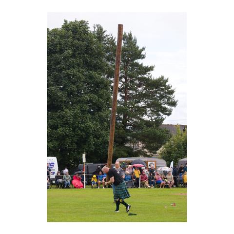 Photo en format portrait. Sur un terrain d'herbe, un homme vêtu d'un kilt porte à la verticale un tronc d'arbre et s'apprête à le lancer. Il est regardé par plusieurs spectateurs.
