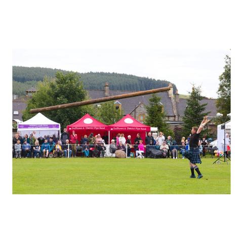 Photo en format paysage. Devant une foule de spectateurs dont certains s'abritent sous des tentes, un homme vêtu d'un kilt vient de lancer un tronc d'arbre. Ce dernier apparaît à l'image en l'air, prêt à retomber plusieurs mètres plus loin.