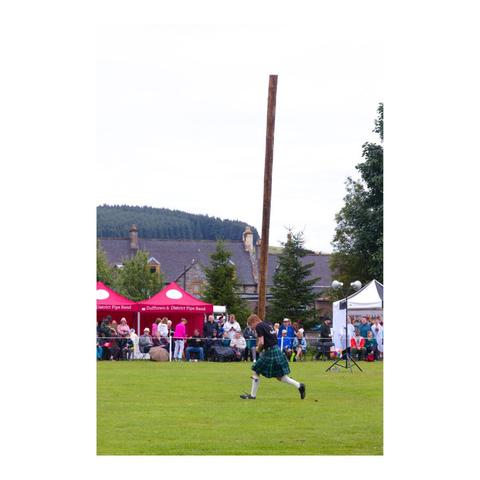 Format portrait. Un homme vêtu d'un kilt porte à la verticale un tronc d'arbre, il est en train de courir afin de lancer le tronc. La scène se passe sur un terrain d'herbe, devant des spectateurs.