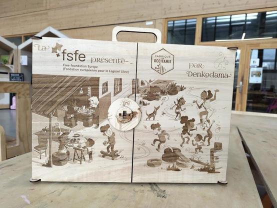 Front of the wooden Kamishibai butai with a scene from the book Ada & Zangemann engraved on the wood, showing the kids tinkering and dancing ; the scene is split on the two doors. The logos of the FSFE, Denkodama and "Fabriqué en Occitanie" are auso displayed. The butai has a handle on the top