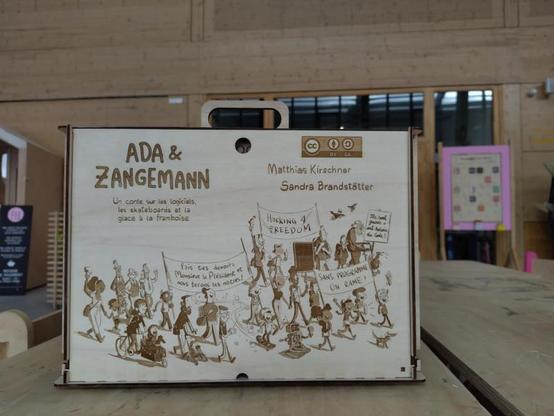 Closed back of the kamishibai butai showing an engraved scene from the book Ada & Zangemann, where kids and adults demonstrate for free, libre open source software. The CC-BY-SA logo is also engraved alongside with the title and the authors names