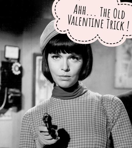 Black and white still from old Get Smart TV series. Female Agent 99 in front of an old public phone pointsba revolver to camera while exclaiming "Ahh... the old Valentine trick !"