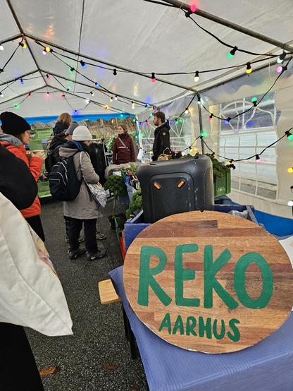People in a tent with windows, coloured lights strung up along the top. A wooden sign with green writing: REKO Aarhus