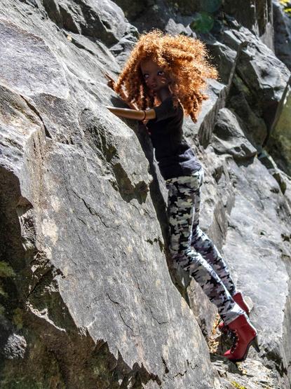 A Smart Doll with curly red hair, a black shirt and camo trousers climbing a rock.