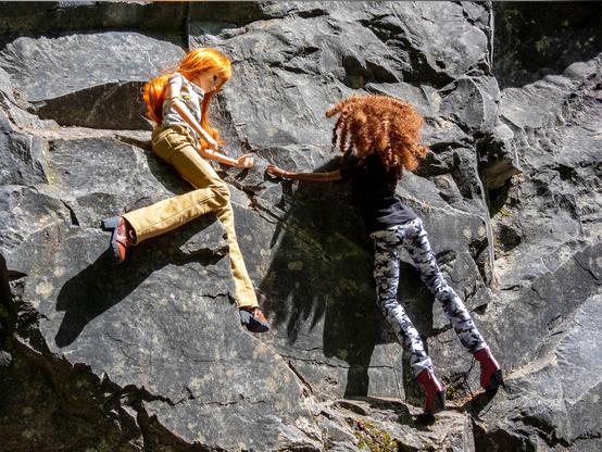 Two Smart Dolls, one with with curly red hair, a black tshirt and camo trousers, one with long red hair, a floral tshirt and corduroi trousers climbing a rock.