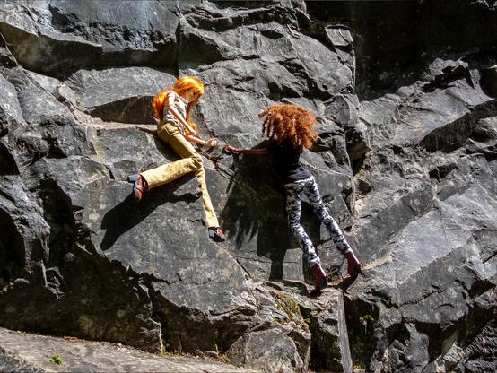 Two Smart Dolls, one with with curly red hair, a black tshirt and camo trousers, one with long red hair, a floral tshirt and corduroi trousers climbing a rock.