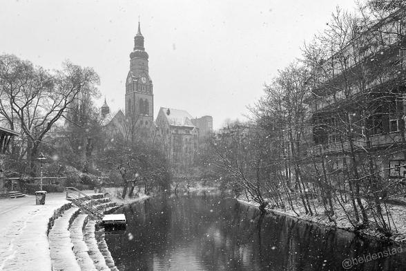 Zwart-wit foto van een winters tafereel, het sneeuwt. We zien een kanaal dat naar naar rechts afbuigt. Links een brede trap die naar het water afdaalt met een kleine steiger. In de achtergrond komt een kerk boven de kale bomen uit.
