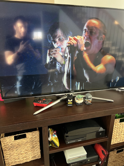 Photo of a TV playing a Nine Inch Nails tour video, showing Trent Reznor and Alessandro Cortini performing a song while my reflection is seen next to them. The TV is on a stand where, several slots below, there is an Xbox 360 E connected to an Xbox 360 HD DVD player. Next to it is the iconic red HD DVD case for the Beside You in Time tour album.