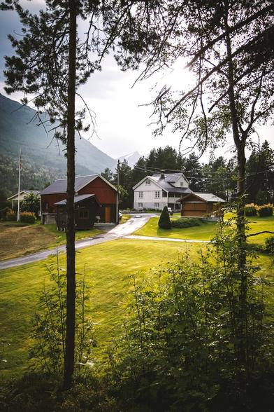 A serene rural scene in Norway with a cluster of houses surrounded by lush greenery and mountain views in the background. A red barn, a white house, and a wooden garage are nestled on a neatly mowed lawn under a soft afternoon light. Trees frame the image, and a flagpole with a Norwegian flag is visible on the left.
