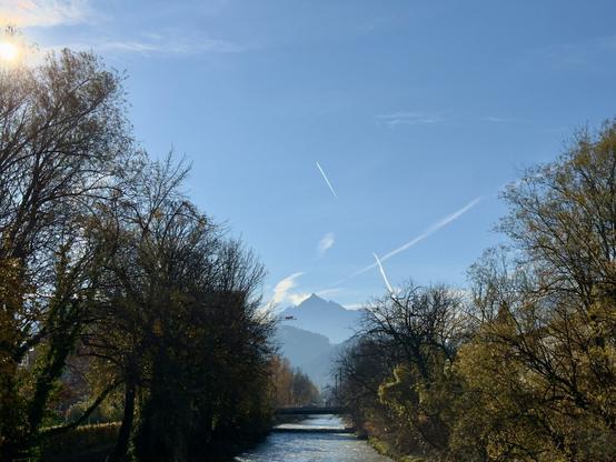 Das Foto zeigt eine malerische Landschaft mit dem Fluss Sill im Vordergrund und dem markanten Berg Serles im Hintergrund. Der Fluss fließt ruhig durch eine von Bäumen gesäumte Umgebung, deren Blätter in herbstlichen Farben leuchten. Eine kleine Brücke überspannt den Fluss, während sich die Silhouette der Serles majestätisch gegen den klaren blauen Himmel erhebt. Die Sonne scheint von links oben ins Bild und erzeugt ein warmes Licht, das die Szene freundlich und einladend wirken lässt. Kondensstreifen am Himmel durchziehen das Bild und verleihen ihm eine dynamische Note. Die Aufnahme vermittelt eine friedliche, fast idyllische Atmosphäre inmitten der Natur.