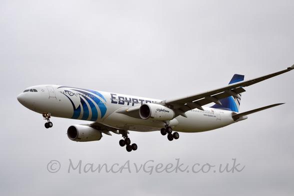 Side view of a white, twin engined jet airliner flying from right to left at a low altitude with it's undercarriage lowered and flaps extended from the rear of the wings, suggesting it is about to land.
There is a large, stylised birds head in shades of black and blue on the forward fuselage, black "EgyptAir" titles on the fuselage over the wings, and swirly black and blue stripes on the tail.
Flat white sky fills the rest of the frame.
