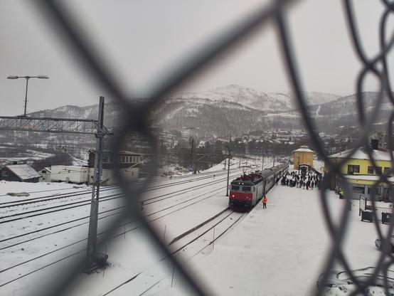 A view from a distance on the train in Narvik. There's a lot of snow and mountains in the background. There's a large group of people on the platform, that just disembarked.