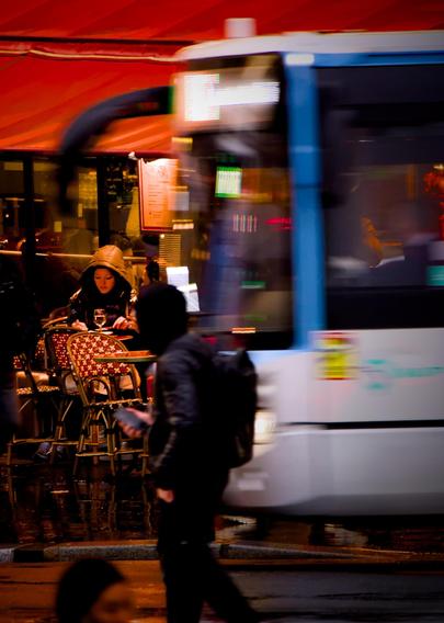 La photographie représente une scène de rue avec un bus au mouvement flou passant devant un café. Une personne est assise à une table à la terrasse du café, sous un auvent rouge, et se concentre sur quelque chose devant elle. L'arrière-plan, l'intérieur du café, est animé et éclairé, ce qui indique qu'il s'agit d'une scène nocturne. Au premier plan, une personne marche en tenant un téléphone à mi-hauteur. Elle est habillée de noir, avec une capuche sur la tête et tourne la tête à l'objectif. L'image dans son ensemble capture une atmosphère urbaine dynamique et animée.