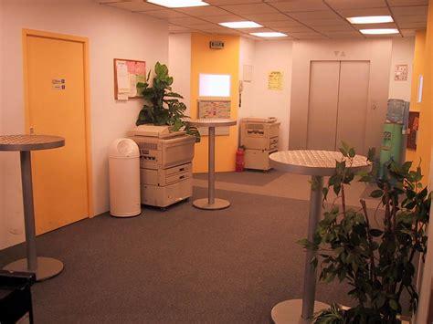 Le décor de la série Caméra Café, une salle de pause dans un espace de bureau, avec de la moquette grise, des murs jaunes, des néons blafards, deux pauvres plantes vertes, une photocopieuse et trois tables hautes en métal.
