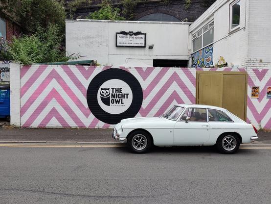 White old MG 2 door coupe in front of a wall with zig zag pink and white lines and a sign for the Night Owl venue. Behind the wall a white nondescript building.