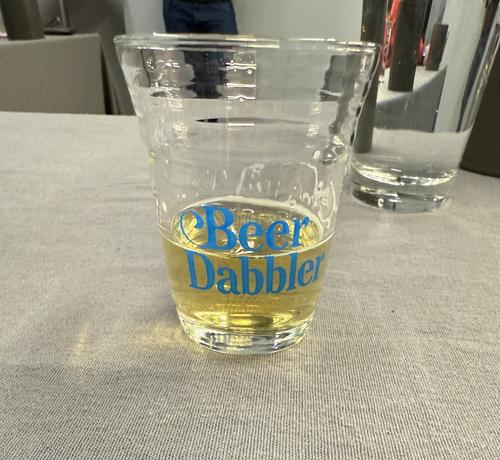 A small glass of light yellow beer. The words “Beer Dabbler” are in light blue on the glass