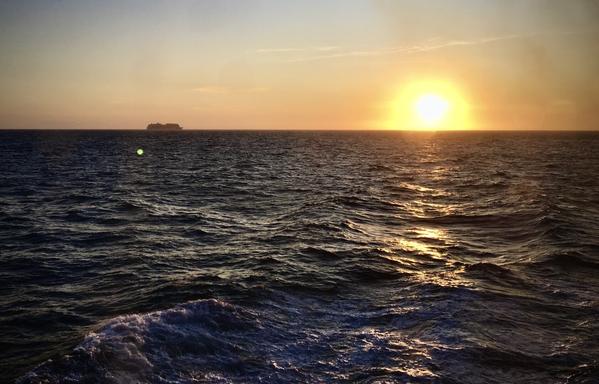 Sunset on the ocean. A ship on the horizon opposite of the sun, also on the horizon.
