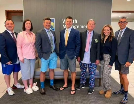 Seven people standing in a row, men & women, at their workplace who’s been asked to come to work wearing the clothes they wear in their zoom meetings. They’re all dressed properly the upper part of the body with a blouse or jacket and shirt while the lower part is a mixture of pyjamas, shorts, and a mix of not coordinated garments in colors that don't match. The image humorously highlights how many people dressed during remote work—formal on top for video calls and casual (or even silly) on the bottom.