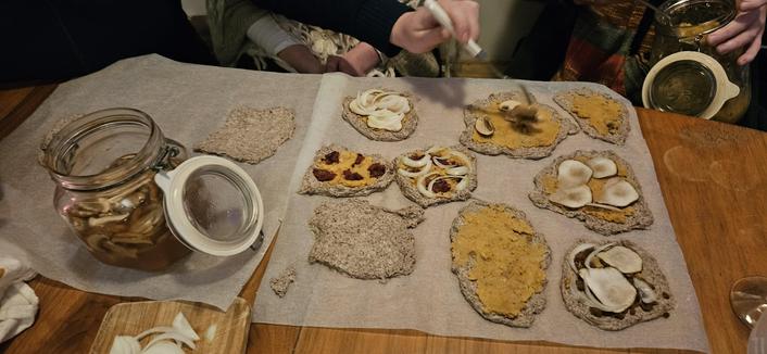 An array of uncooked sourdough purple wheat mini pizzas with fermented red lentil paste, beet/bean/miso frikadeller crumbled on top, mushroom slices fermented in brine, onion slices, sunchoke slices. A jar on the side. Hands with a jar in the background.