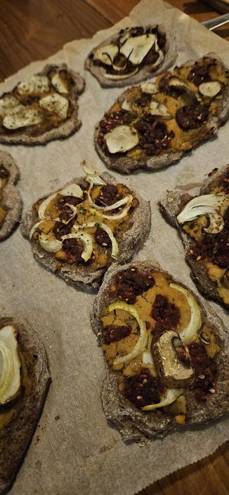 An array of sourdough purple wheat mini pizzas with fermented red lentil paste, beet/bean/miso frikadeller crumbled on top, mushroom slices fermented in brine, onion slices, sunchoke slices.