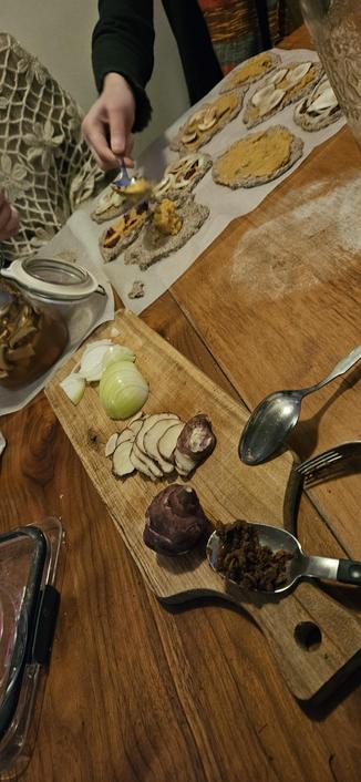 A wooden cutting board with slices onion, sunchoke, red beet & bean patty. In the background a hand places red lentil fermented paste on a mini pizza.