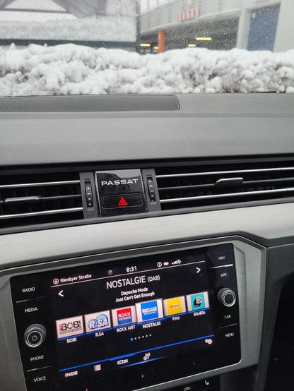 Das Bild zeigt das Innere eines Autos, wahrscheinlich eines VW Passat, aufgenommen vom Fahrersitz aus. Das Armaturenbrett ist sichtbar, mit Lüftungsschlitzen, dem Schriftzug "Passat" und einem Warnblinklichtschalter. Der Radiodisplay zeigt den DAB-Radiosender "Nostalgie" an, der gerade "Depeche Mode - Just Can't Get Enough" spielt. Vor der Windschutzscheibe liegt eine dicke Schicht Schnee. Die Perspektive ist leicht nach unten gerichtet.