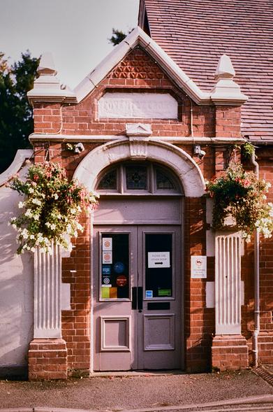 A rather ornate (ex) doorway with pillars and fancy brickwork, with a little peaked gable. There are hanging baskets each side of the door. The glass panels in the door have many stickers on them with various awards, including Michelin 2021. Other signs say "No deliveries to this door" and "Please use other door". Portrait format colour photo.