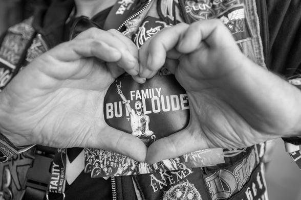 Schwarzweißfotografie, Detailbild. EIne Person in Kutte zeigt mit den Händen eine Herzform. Mit dieser Geste hält die Person eine Postkare an  seine Kutte, sodass in dem Herz der Metality-Slogan "Family. But Louder." zu erkennen ist.
