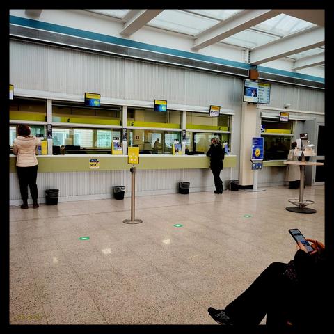 Quadratisches Farbfoto mit schwarzem Rahmen, aufgenommen in einem Postamt in Rom. In Bildmitte drei belegte Schalter, dazwischen zwei ohne Personal. Im Vordergrund rechts ein Teil einer wartenden Person mit Telefon in der Hand. Oben ein Glasdach, darunter weiß gestrichene Eisenträger. An einer Säule darunter angebracht ein Display mit Nummern.