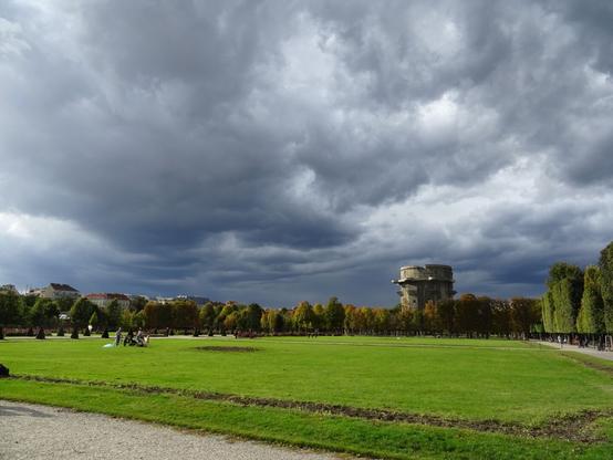 Das Bild zeigt einen weitläufigen Park mit einer großen, grünen Rasenfläche im Vordergrund und einem breiten Weg im unteren Bereich. Im Hintergrund sind Bäume zu sehen, die herbstlich gefärbt sind, sowie vereinzelte Gebäude. Dominant rechts im Bild steht ein massiver Flakturm aus Beton, ein Relikt aus dem Zweiten Weltkrieg. Über dem Park ziehen dunkle, dramatische Wolken auf, die eine bedrohliche Stimmung erzeugen und auf ein nahendes Gewitter hindeuten. Einzelne Spaziergänger und Radfahrer sind im Park unterwegs, was eine ruhige, aber angespannte Atmosphäre widerspiegelt.