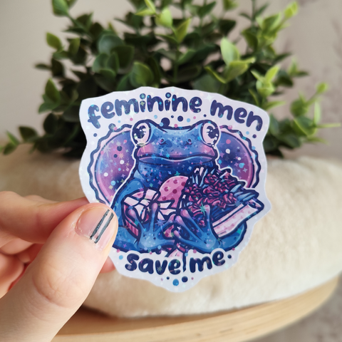Hand holding up a sticker of a frog carrying a box of chocolates and a bouquet of flowers, titled "feminine men, save me"