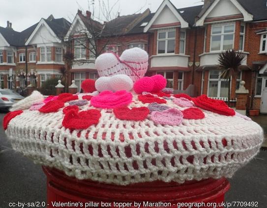 The top of a UK red postbox, other the circular pillar box style.  It has been decorated with a crochet net of cream wool, on which are sewn crochet red and pink rose petals and love hearts