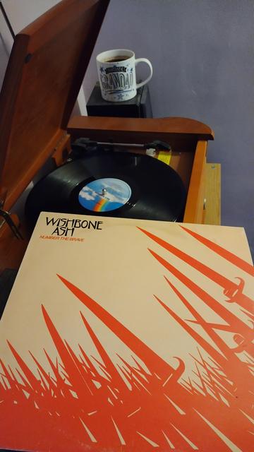 Record player with a 12" black vinyl record.
In the foreground is an album cover showing a mass of red arms holding red swords held up against a creamy  yellow background.
Behind the player is a mug of coffee on a speaker.