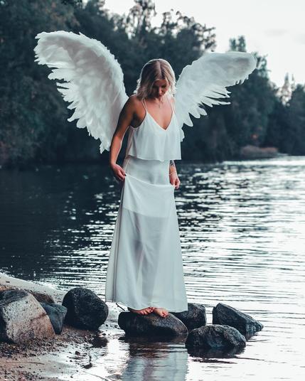An ethereal portrait of a model with white angel wings standing on rocks by the river, wearing a flowing white dress, captured by Kuvaajankulma.