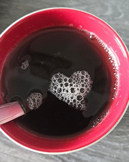 Photo looking down into a red mug filled with a dark liquid (tea in this case). A spoon leans agains the edge. Some tiny white foam bubbles shape a delicate heart on the surface 💜