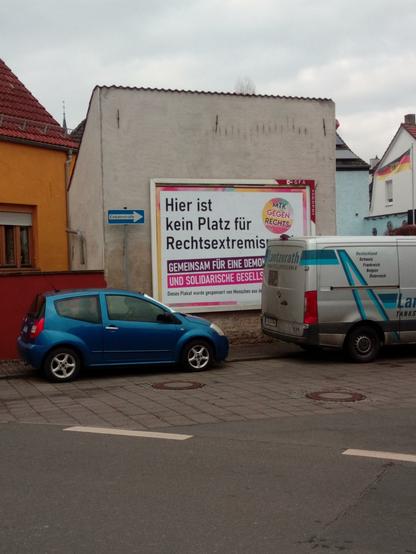 an einer Hauswand hängt ein Plakat mit der Aufschrift "Hier ist kein Platz für Rechtsextremismus. Gemeinsam für eine demokratische und solidarische Gesellschaft." Davor zwei Autos.