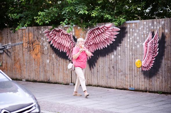 An elderly woman in a pink shirt walks along the sidewalk, an ice cream cone held in her hands. Behind her, a weathered wooden fence serves as a canvas for a graffiti of vibrant pink angel wings, lining up perfectly with her back.