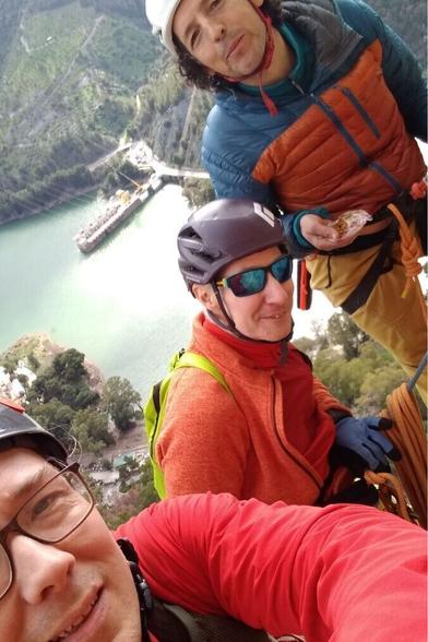 Selfie of 3 men in climbing helmets and gear posing at a belay station high on a cliff with a reservior far below in the background