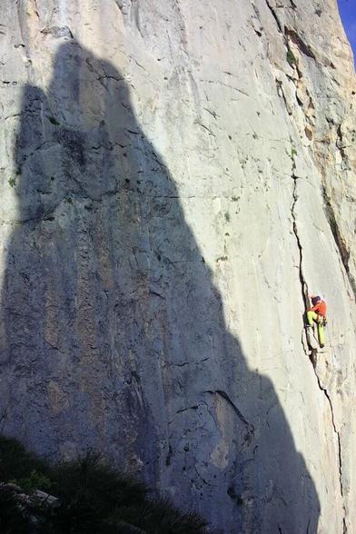 A grey, limestone cliff face with a sharp shadow outline diagonally across. A brightly-clad climber moving up an obvious flake halfway up right of frame