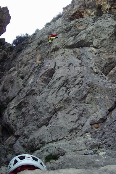 A grey limestone face, steep with a brightly-clad climber moving over a bulge of rock