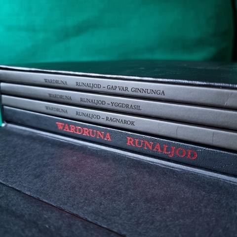 Opened lid of the "Vinyl Deluxe Boxset" shows three matte black Gatefold spines that are printed with UV varnish, that read:
1) WARDRUNA RUNALJOD - GAP VAR GINNUNGA
2) WARDRUNA RUNALJOD - YGGDRASIL
3) WARDRUNA RUNALJOD - RAGNAROK

Also there is a same sized spine of a book that has the same black faux leather material with red silverfoil printing, which reads:
WARDRUNA RUNALJOD